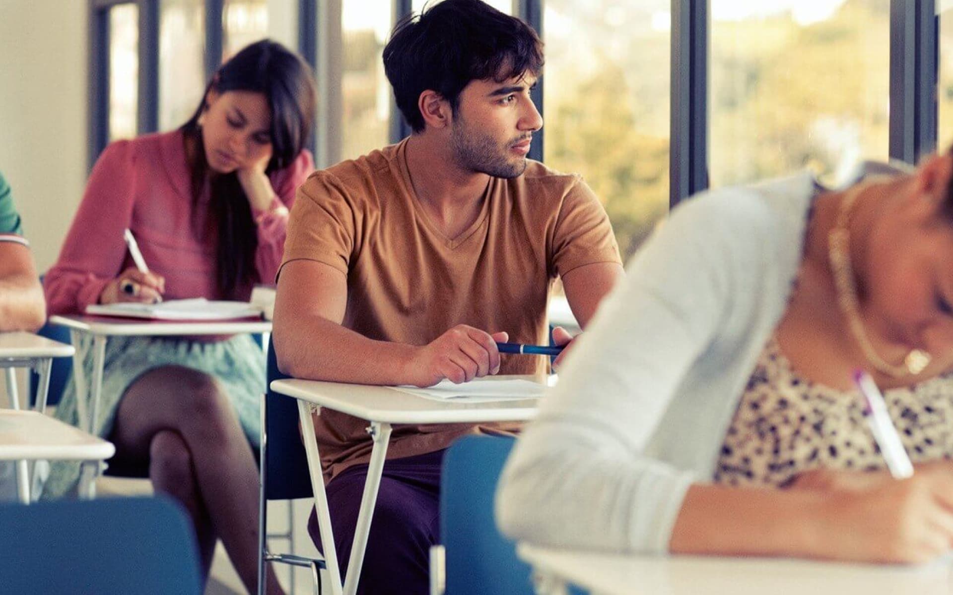 Several students are seated at desks in a classroom setting, diligently writing. The man in the center gazes thoughtfully to the side, while others focus on their tasks.