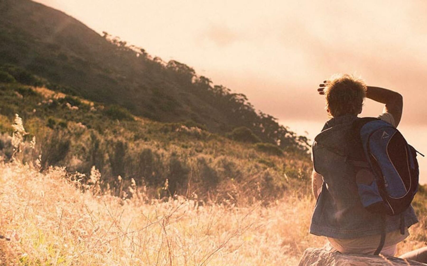 A person with a backpack is looking out over a vast, sun-drenched landscape with rolling hills and dry grass, suggesting a sense of adventure and exploration.