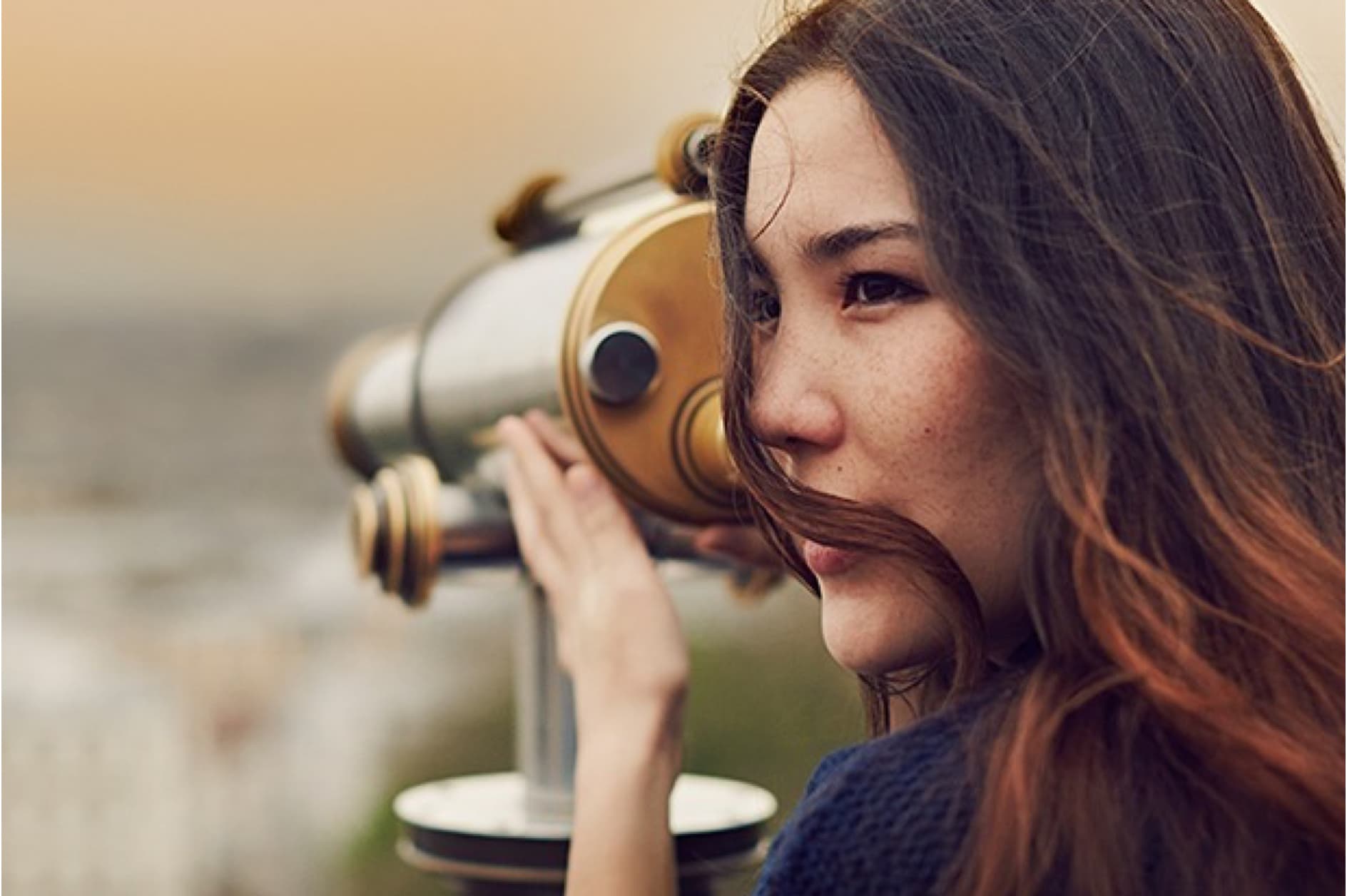 A young woman with long brown hair looks through a telescope into the distance as the wind blows her hair. She appears to be enjoying a view.