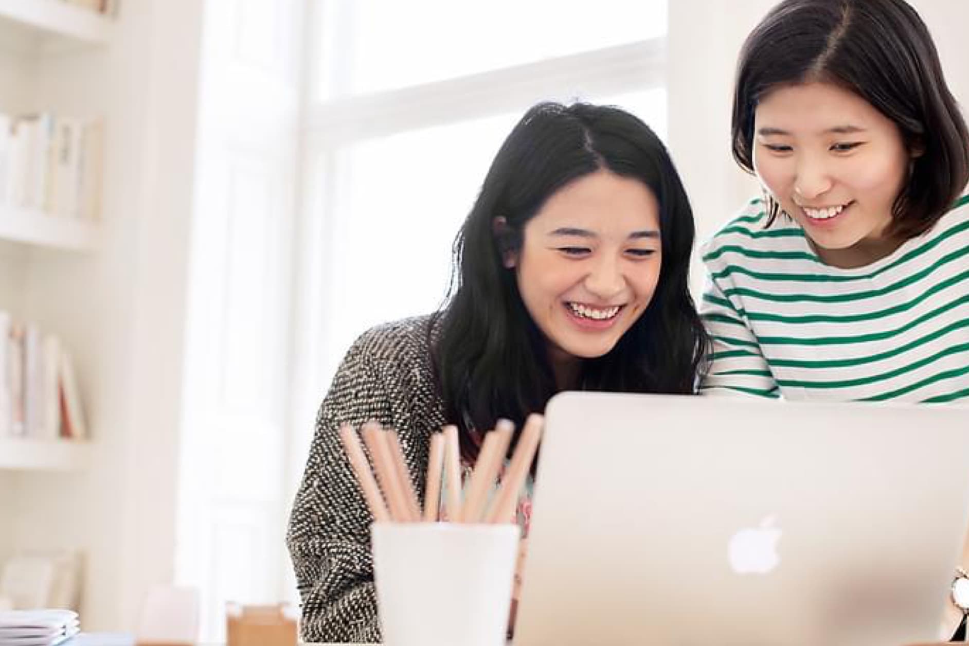 Two young Asian women are looking at a laptop with smiles, appearing engaged and happy with what they are seeing on the screen. They are in a bright, modern setting.