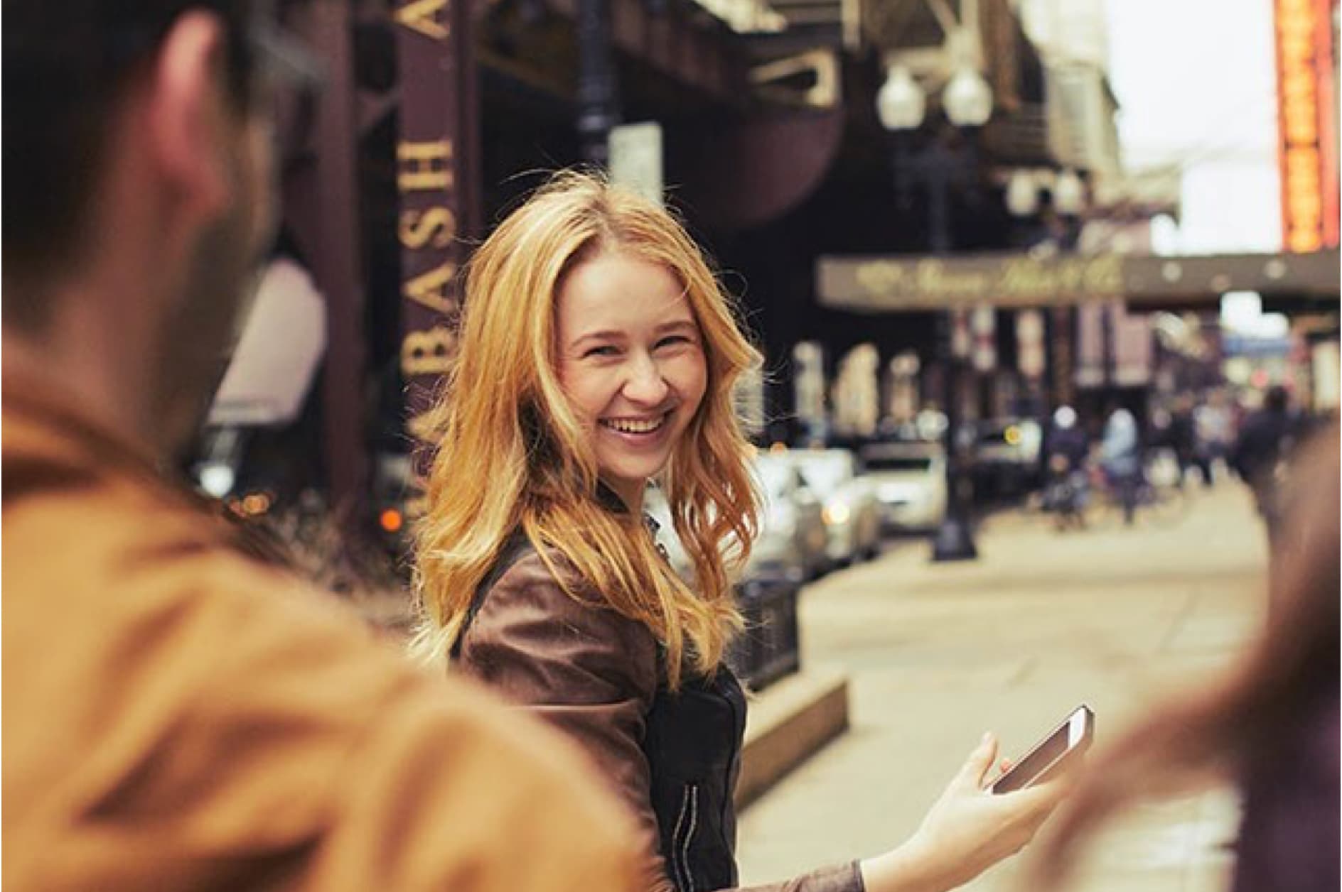 A young blonde woman smiles and holds a smartphone while walking down a busy city street. She is looking over her shoulder at the camera.