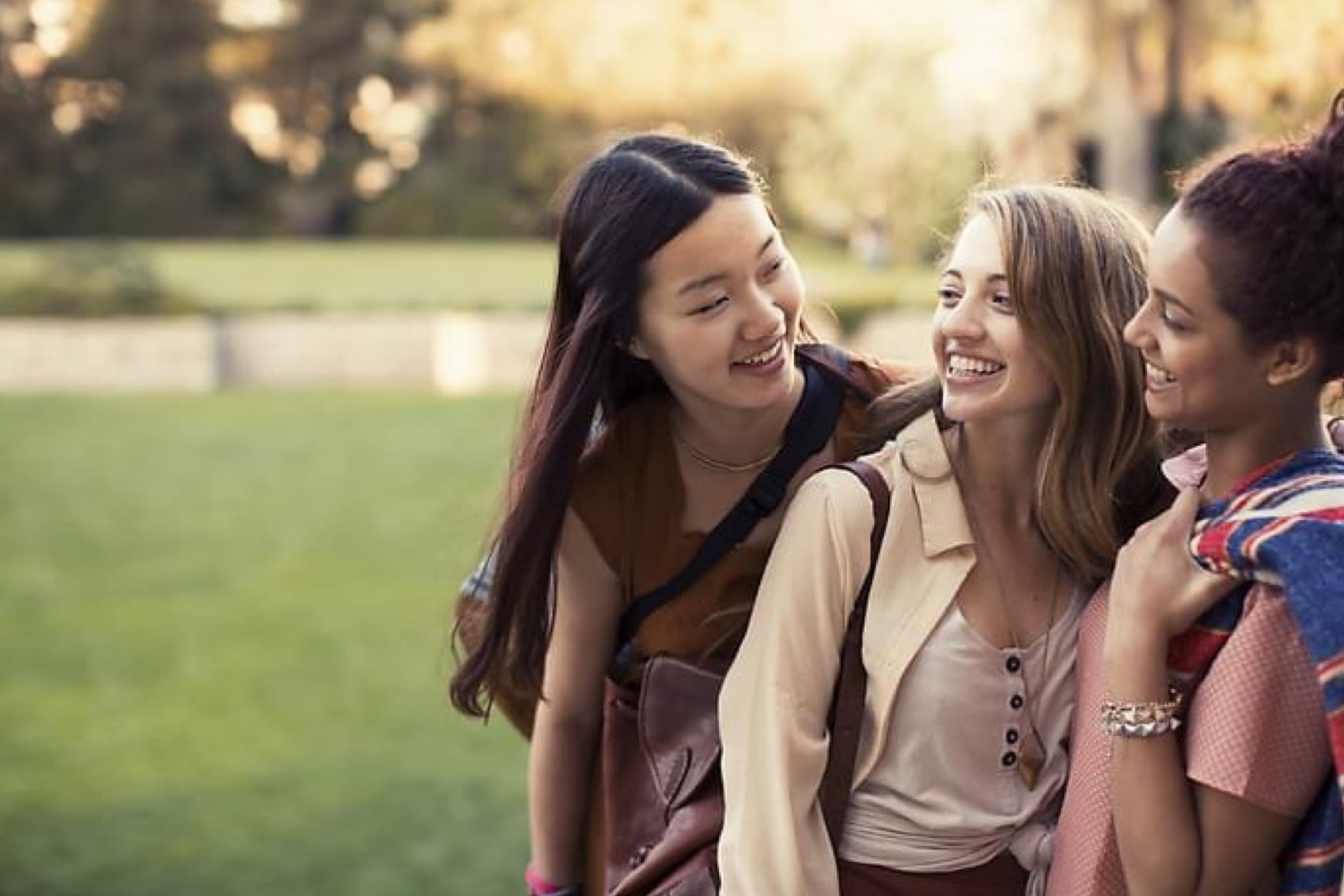Three young women of diverse ethnicities are laughing and conversing outdoors as the sun shines through the trees.