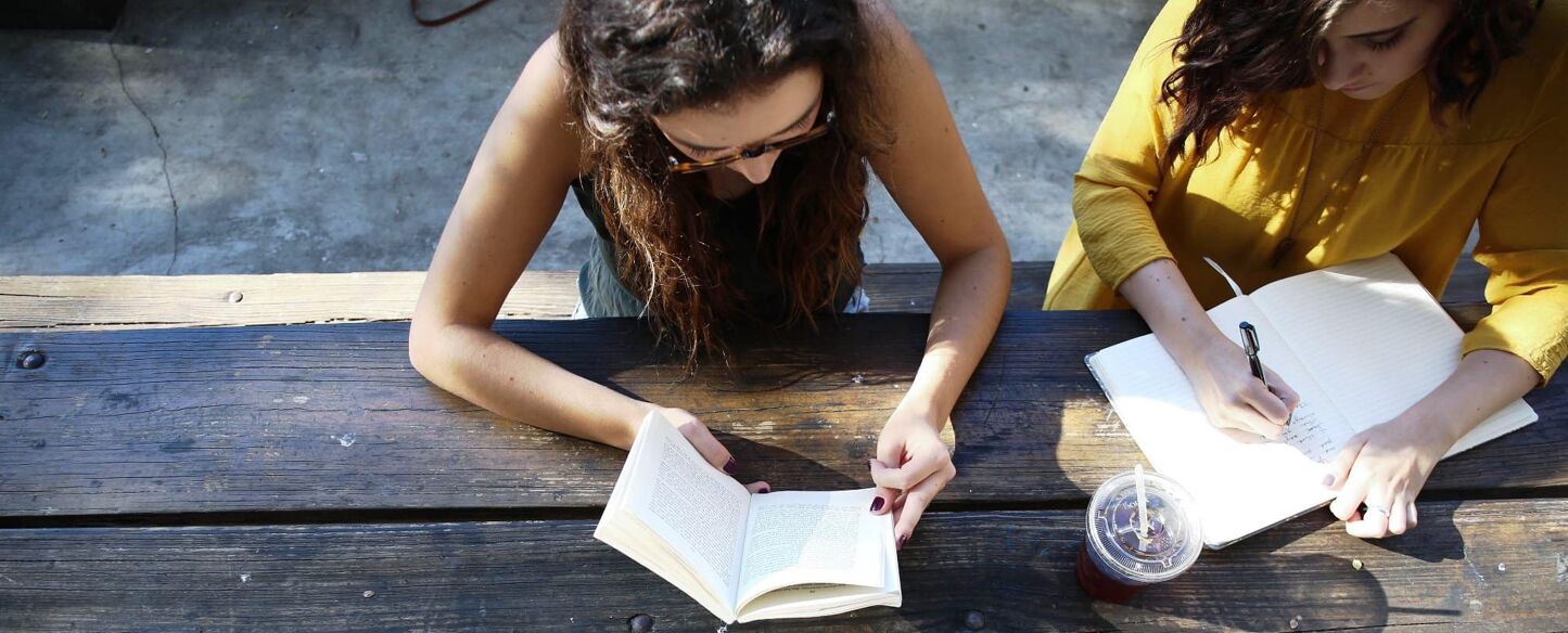 Two young women are sitting at an outdoor wooden table. One is reading a book while the other is writing in a notebook.