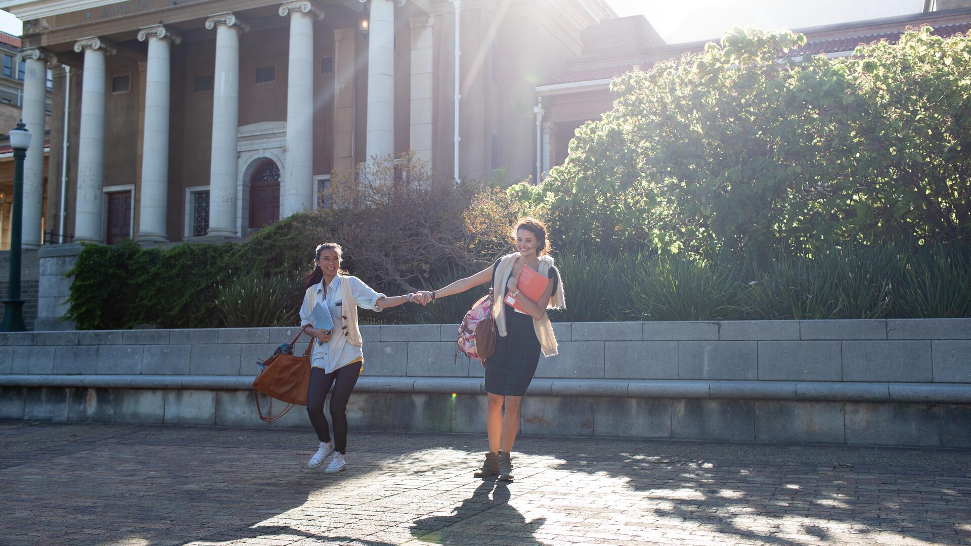 Two young women are walking hand-in-hand on a sunny campus in front of a historic building with columns. They are carrying books and bags, suggesting an academic setting.