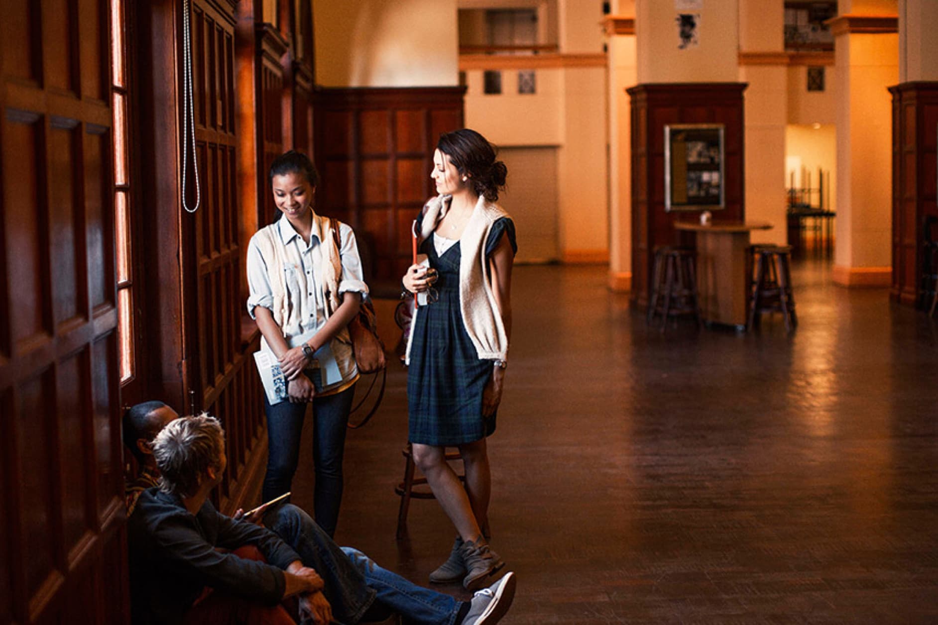 Three people are in a large, wood-paneled room with a polished wooden floor. Two women are standing and talking near a window, while a third person sits on a bench.