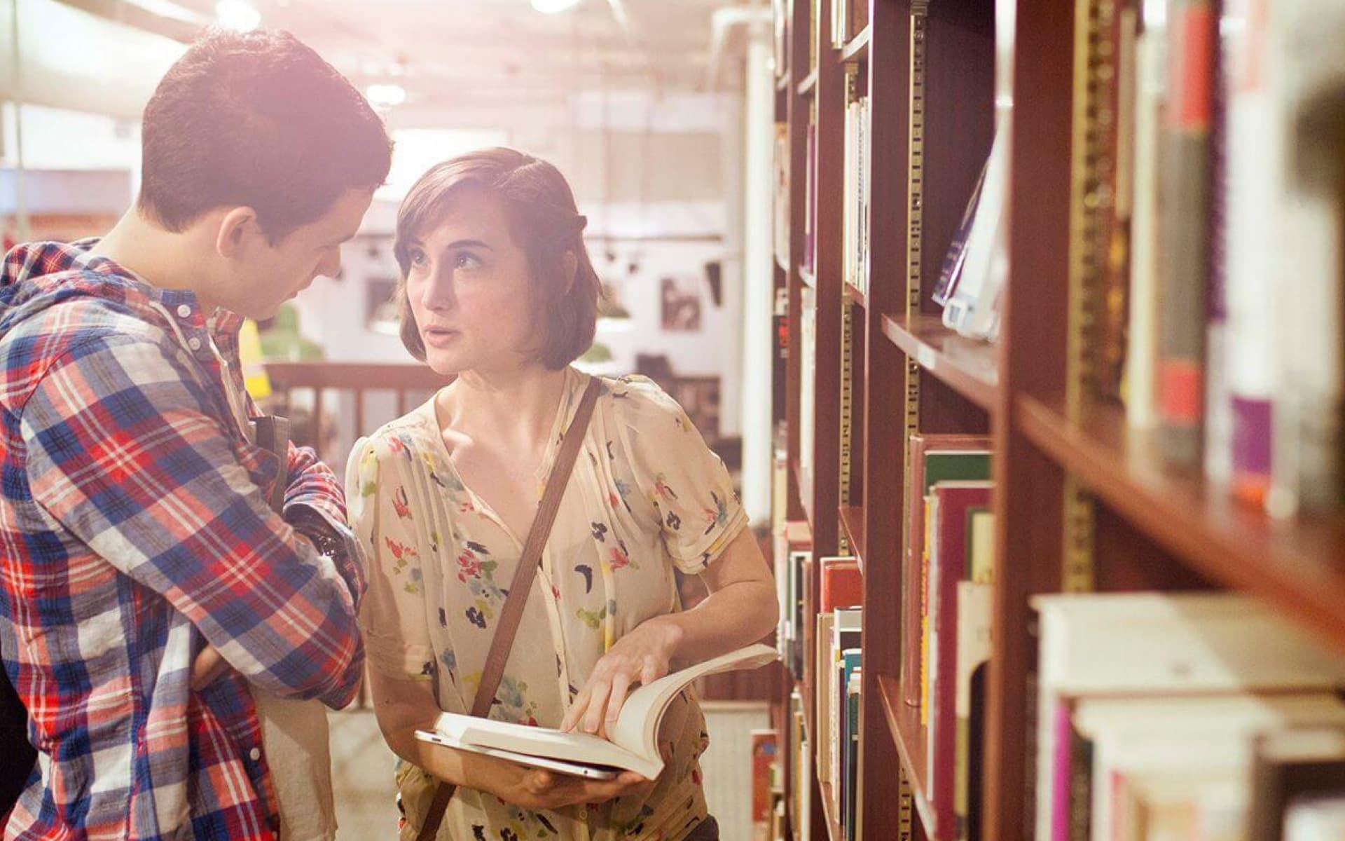 Two young adults are browsing books in a library aisle, with one holding an open book and appearing to ask a question.