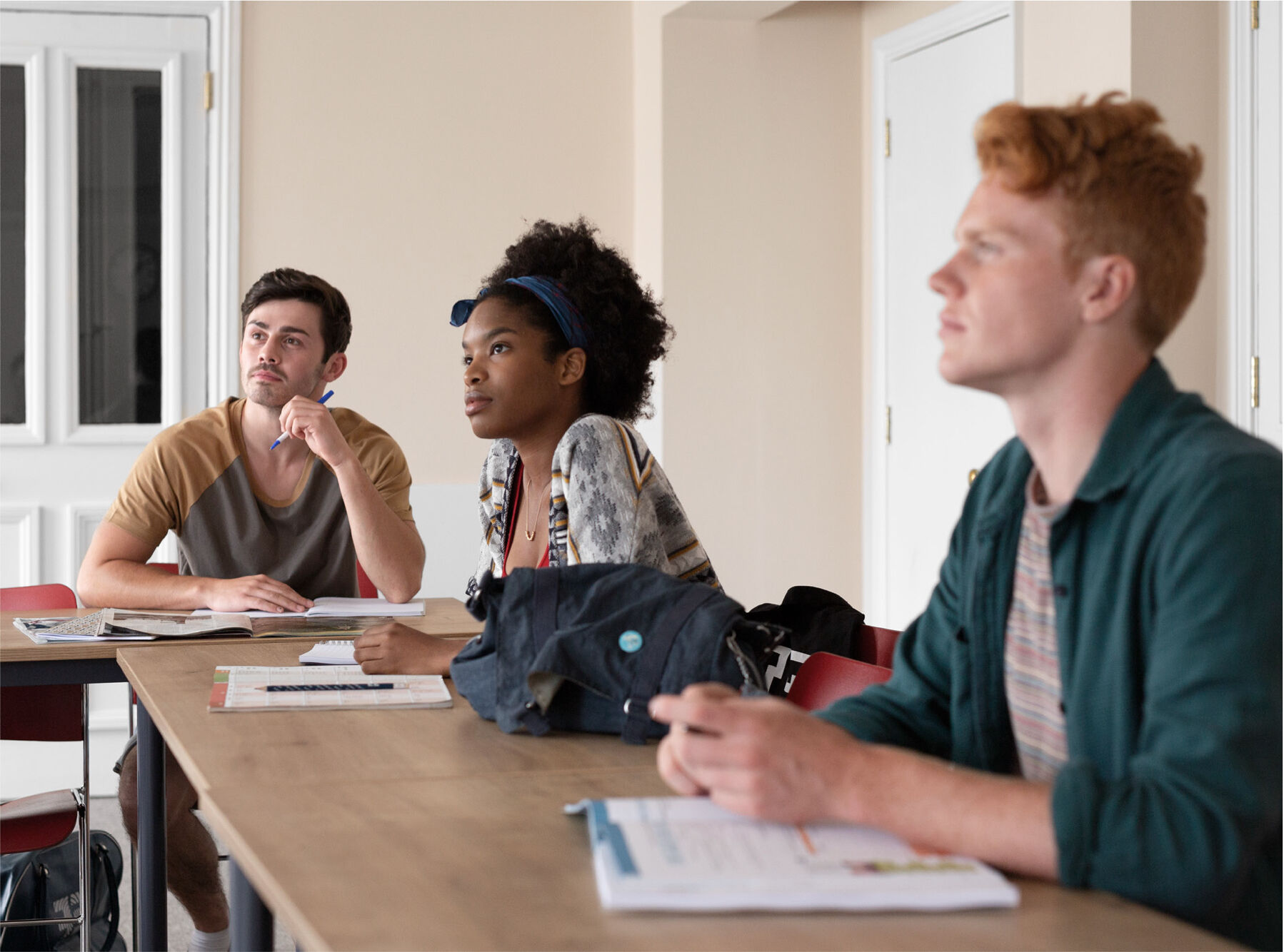 Three students, two men and one woman, are sitting at desks in a classroom, attentively listening to a lesson. They appear engaged in the educational setting.