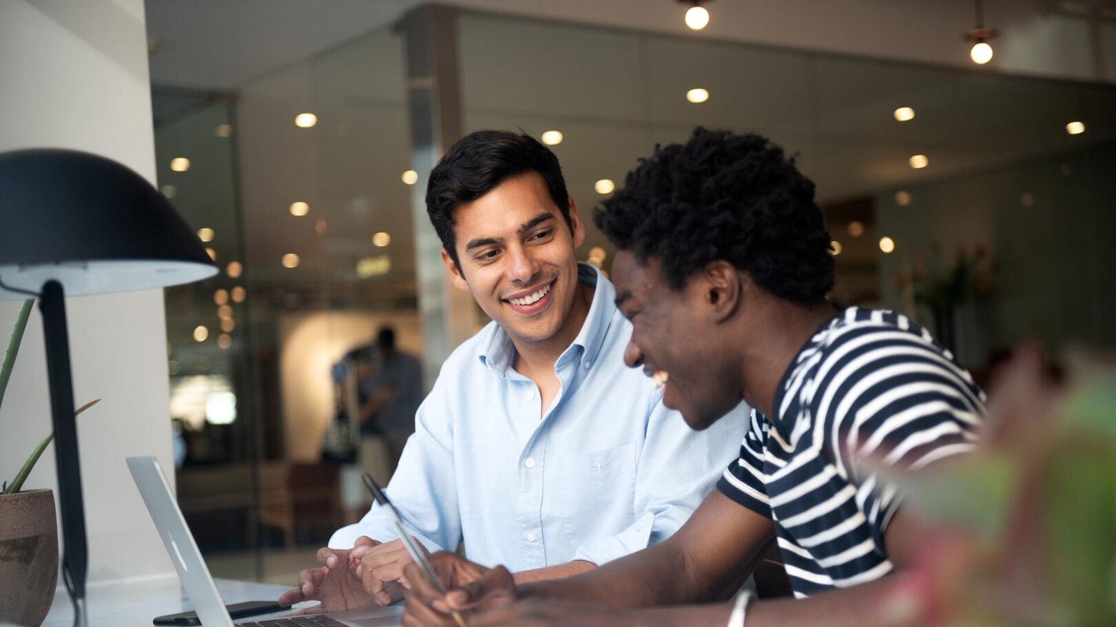 Two young men, one in a light blue shirt and the other in a striped shirt, are smiling and looking at a laptop while collaborating.