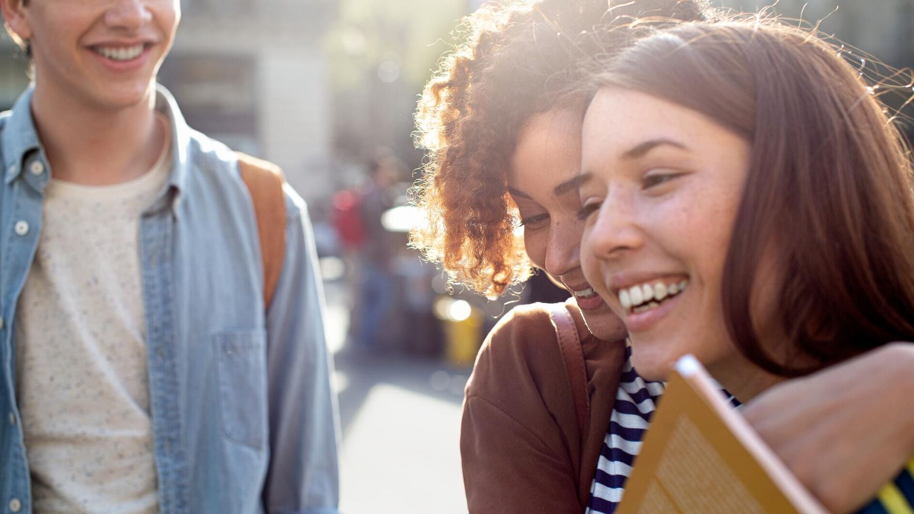Three young people are laughing and walking down a street as the sun shines. They appear to be friends, enjoying their time together.