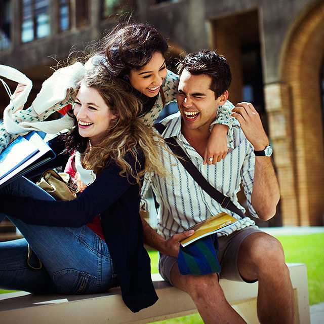 Three laughing students are sitting on a bench outside a building, enjoying the sun and holding books and bags.