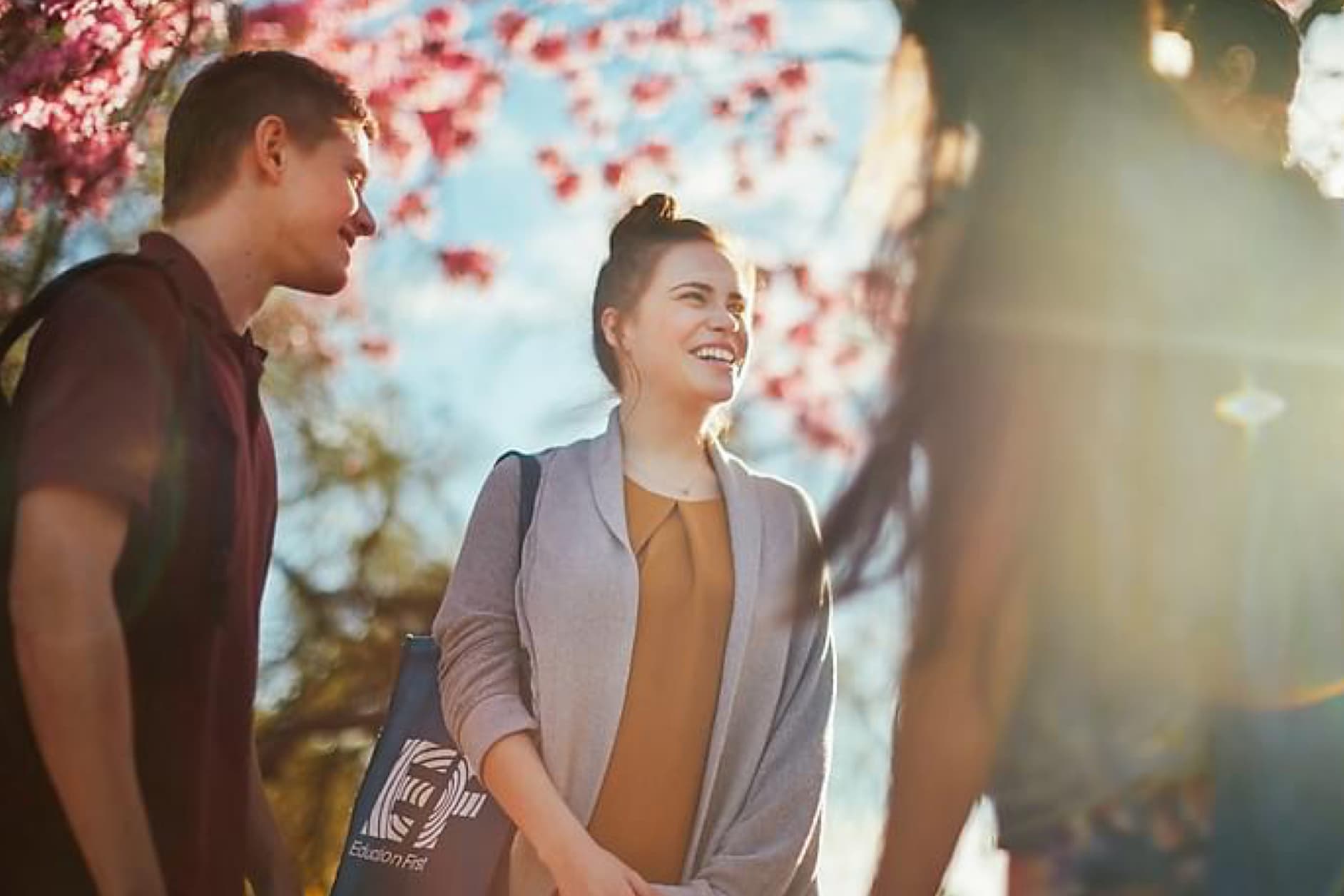 Three young students are walking outdoors on a sunny day under blooming cherry trees. They are smiling and engaged in conversation while carrying their backpacks.