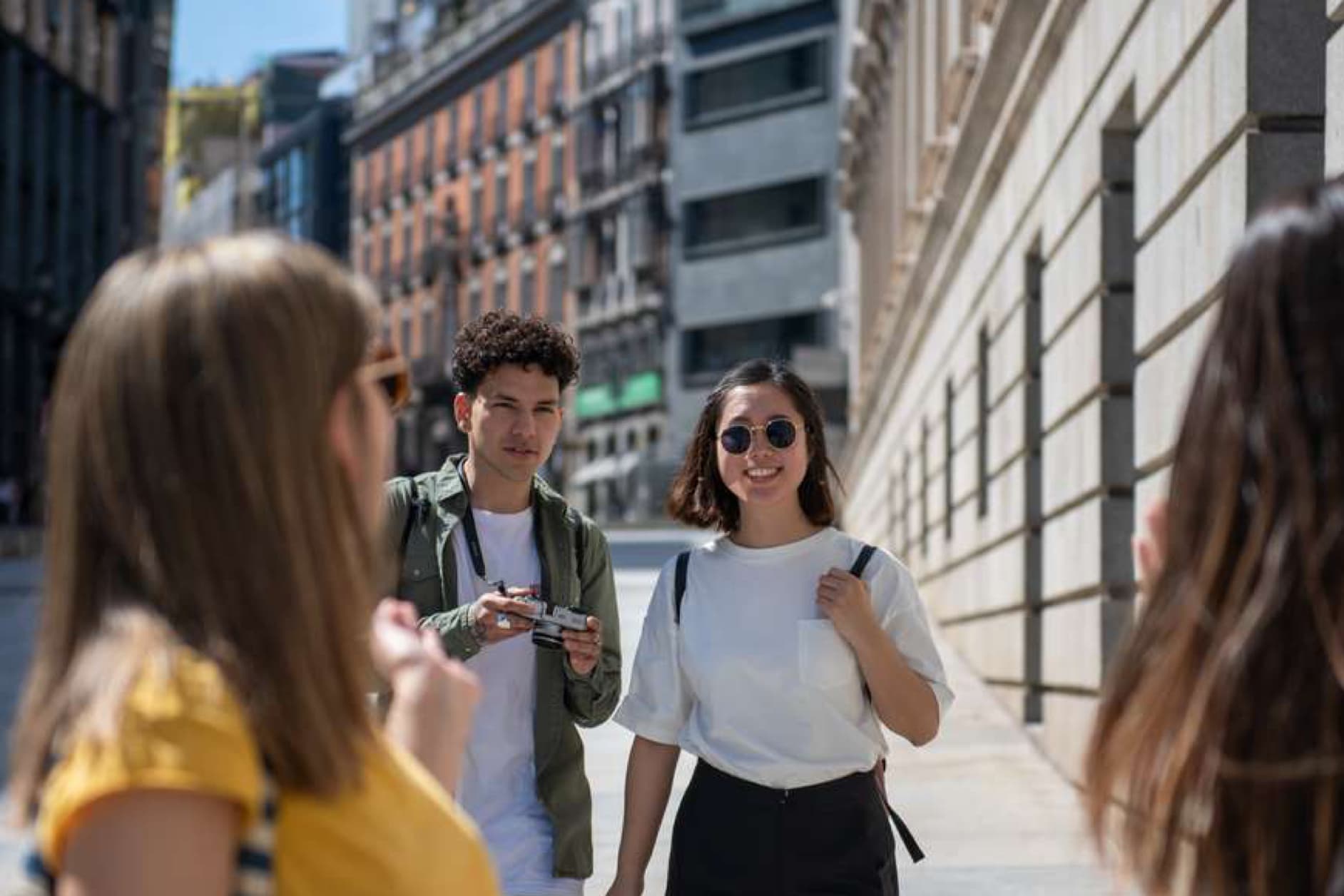 A group of four young friends are walking down a sunny city street, with two in the foreground and two in the background. One of the men is holding a vintage camera.