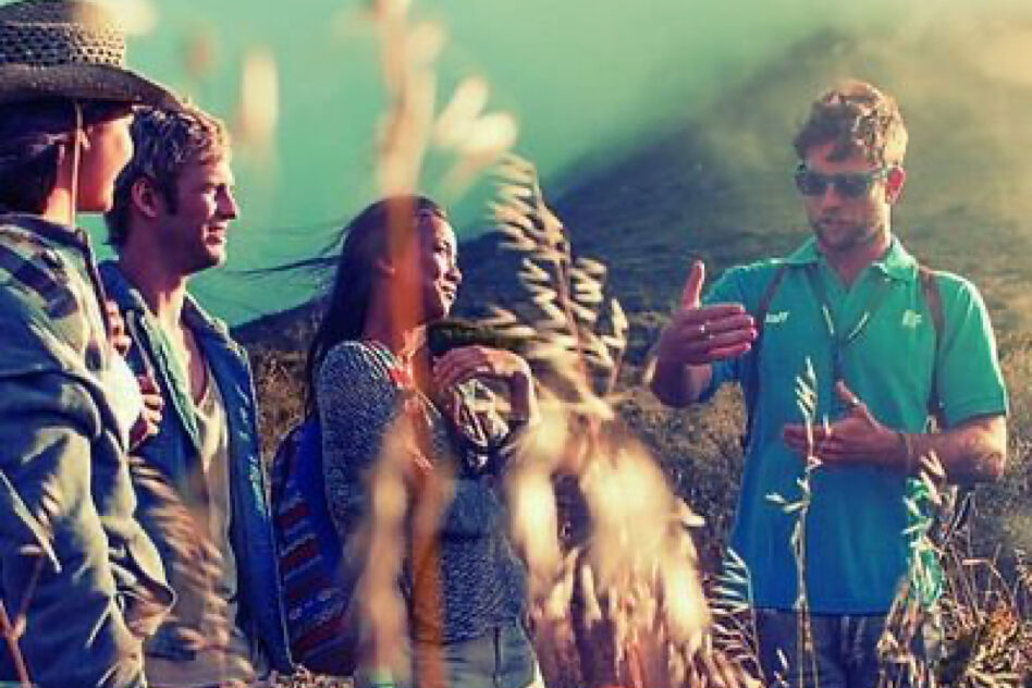 A tour guide is enthusiastically explaining something to a small group of tourists while gesturing with his hands in a scenic, mountainous outdoor setting.