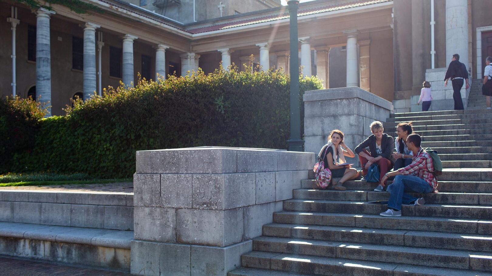 A group of young adults are casually sitting and talking on the wide stone steps of a classical-style building with large columns, enjoying a sunny day outdoors.