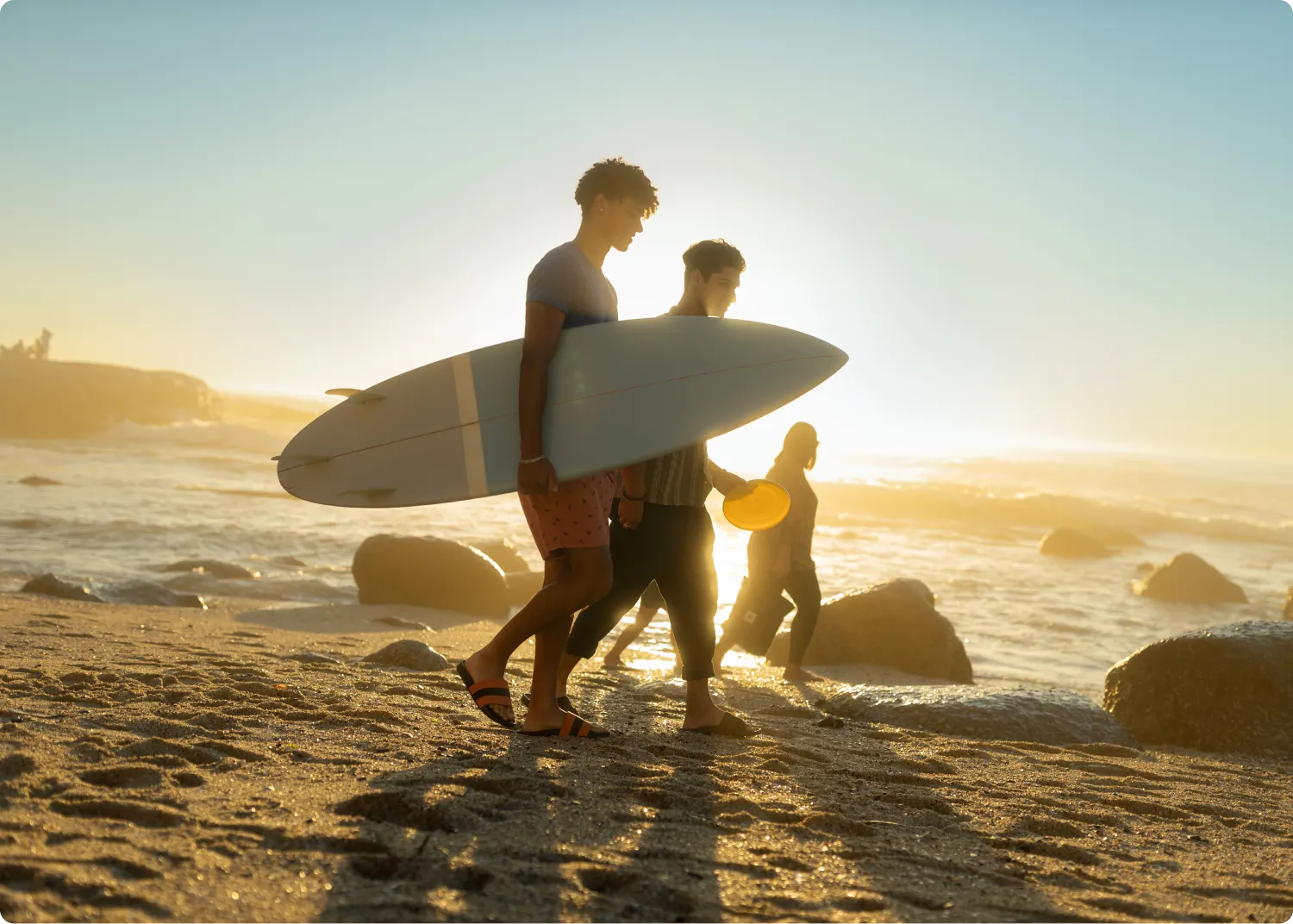 Two young men walk along a sandy beach at sunset, carrying a surfboard. The sun